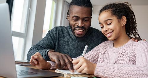 Image of a young student in a pink sweater taking an online class with her learning coach dad who is wearing a grey sweater. 